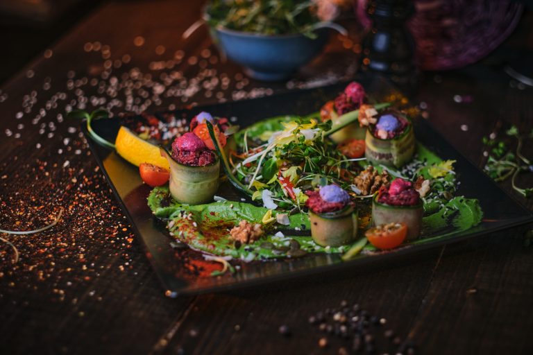 Rollitos de pepino rellenos, ensalada de hojas verdes, tomates cherry, nueces y flores comestibles sobre un plato oscuro.