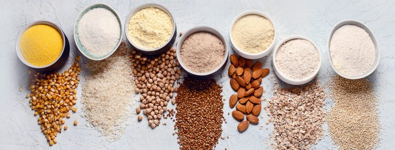 Fila de cuencos con harinas de varios colores sobre granos, legumbres, almendras y copos de avena.