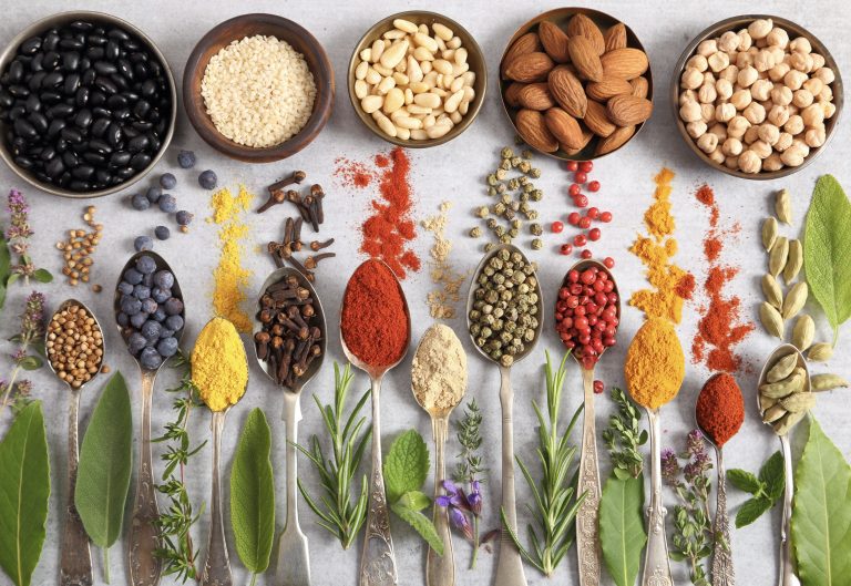 Cuencos de legumbres, frutos secos y cereales junto a cucharas con especias de colores y hierbas frescas.