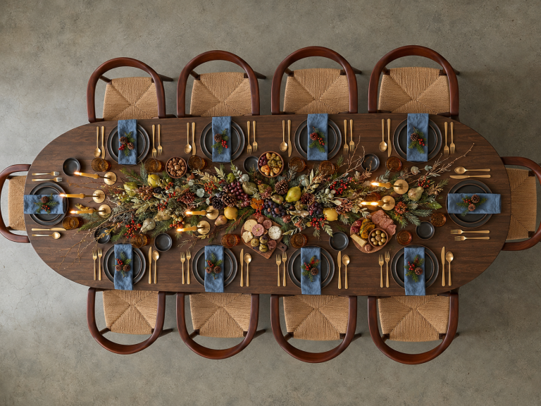 Vista cenital de una mesa de evento de madera decorada con un centro botánico continuo de ramas, piñas, uvas y peras, con platos grises, servilletas azules y candelabros.