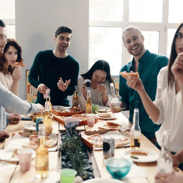 Grupo de amigos compartiendo pizza alrededor de una mesa larga en el interior.